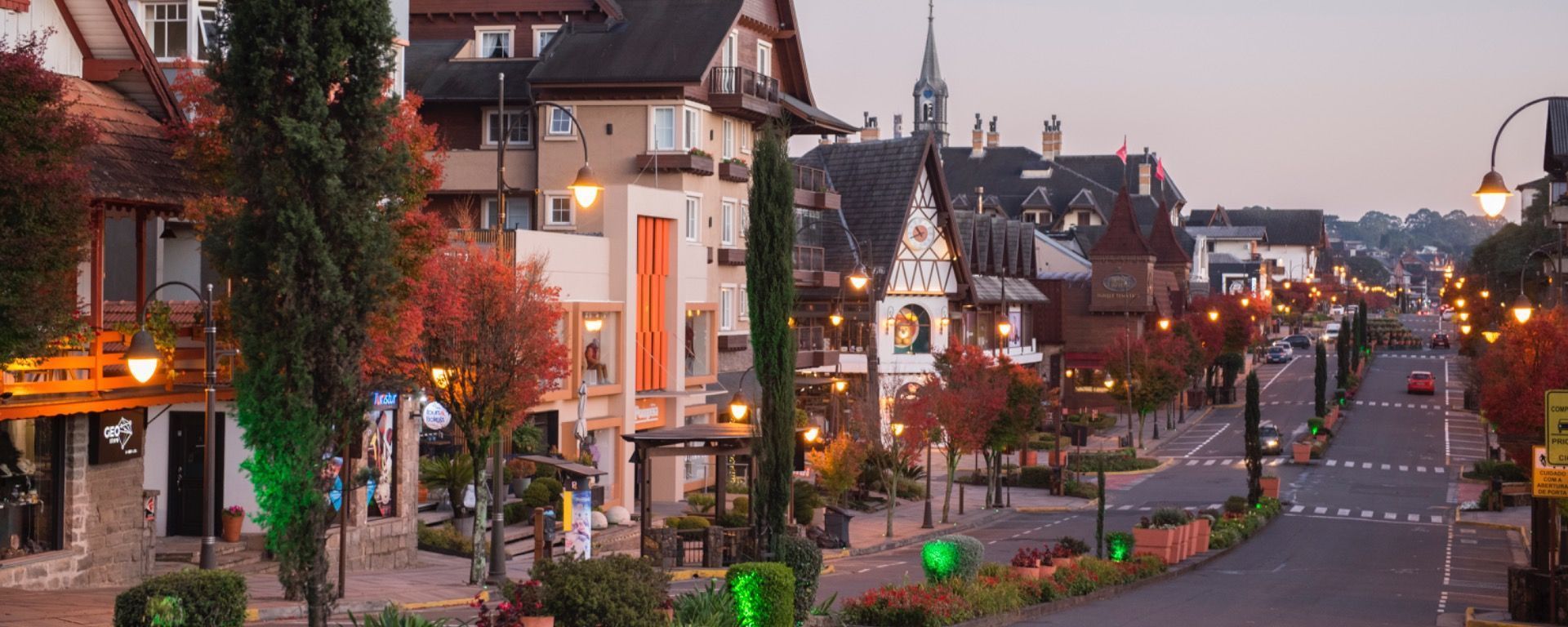 Rua ao entardecer em uma cidade europeia, com prédios iluminados, árvores e uma rua tranquila ladeada por canteiros de flores.
