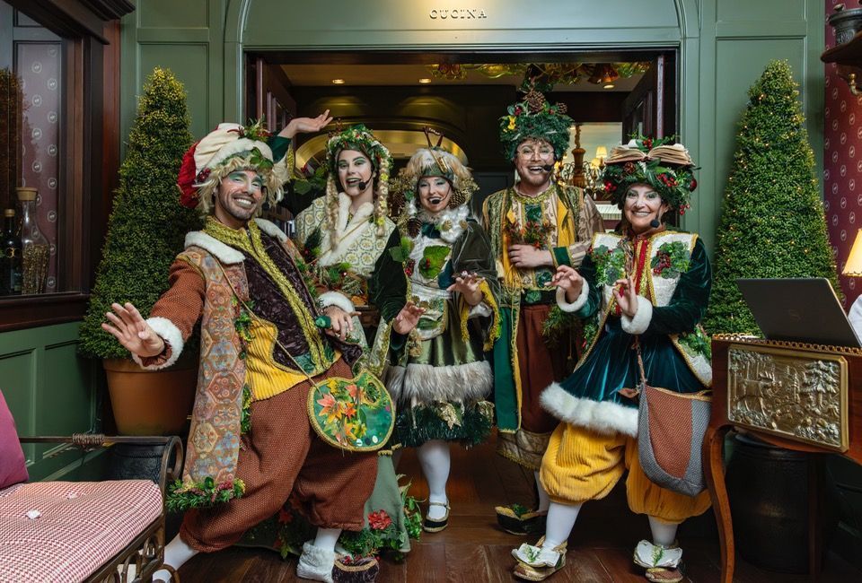 Cinco artistas com trajes coloridos posando em frente à entrada de um prédio, sorrindo e segurando adereços.