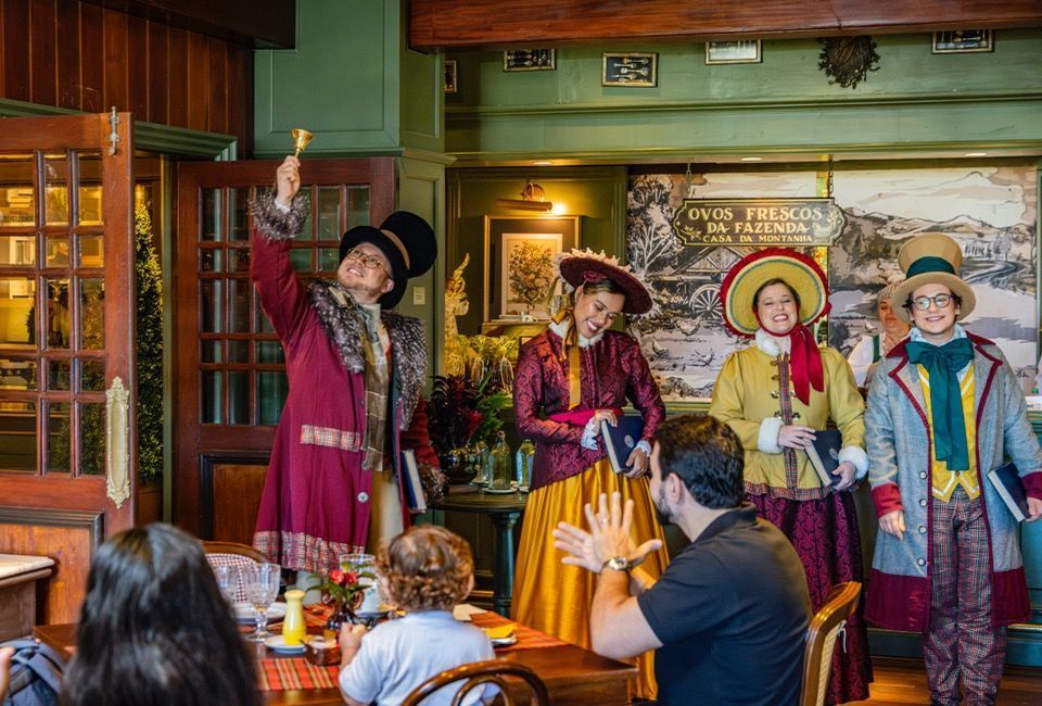 Cinco pessoas com trajes coloridos se apresentam em um pub aconchegante, acenando e cantando para os clientes sentados.
