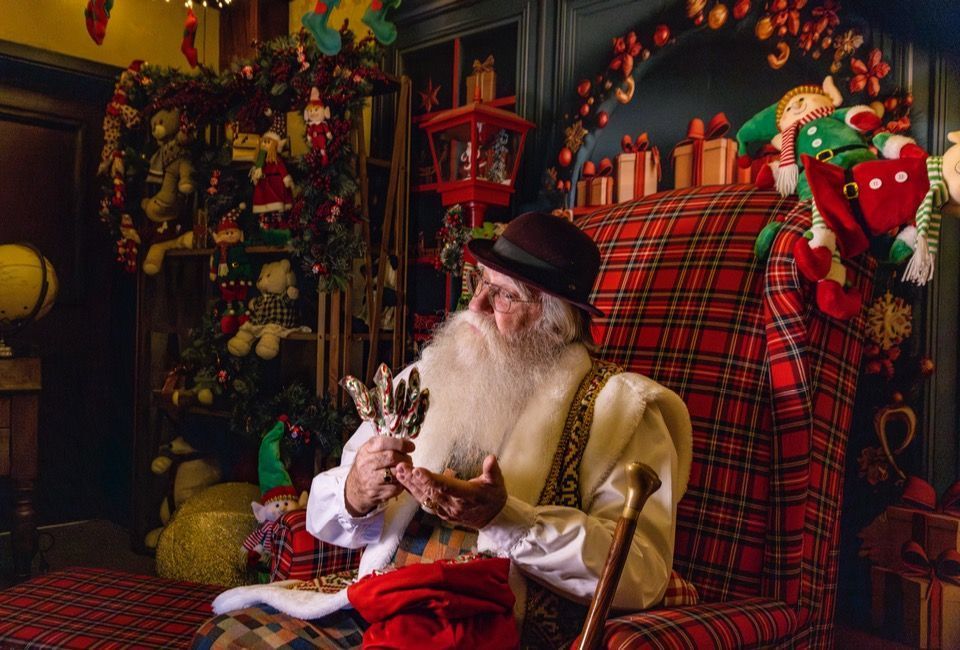 Papai Noel lendo um bilhete em uma oficina aconchegante com uma cadeira xadrez vermelha e decorações festivas.