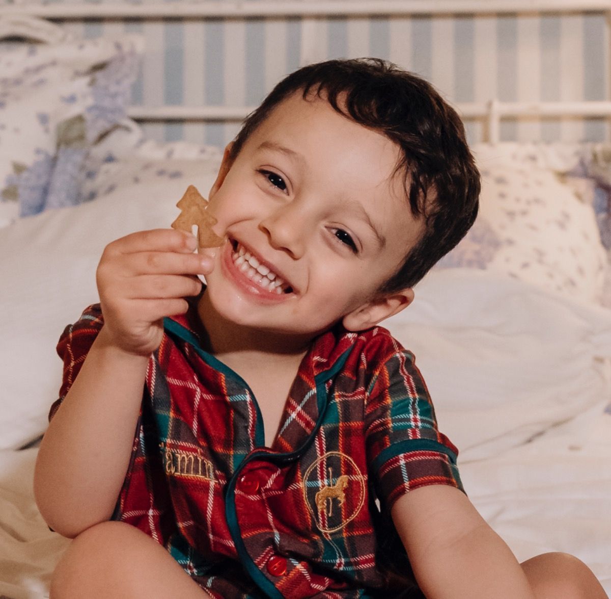Criança sorridente com camisa xadrez vermelha segurando um biscoito em uma cama.