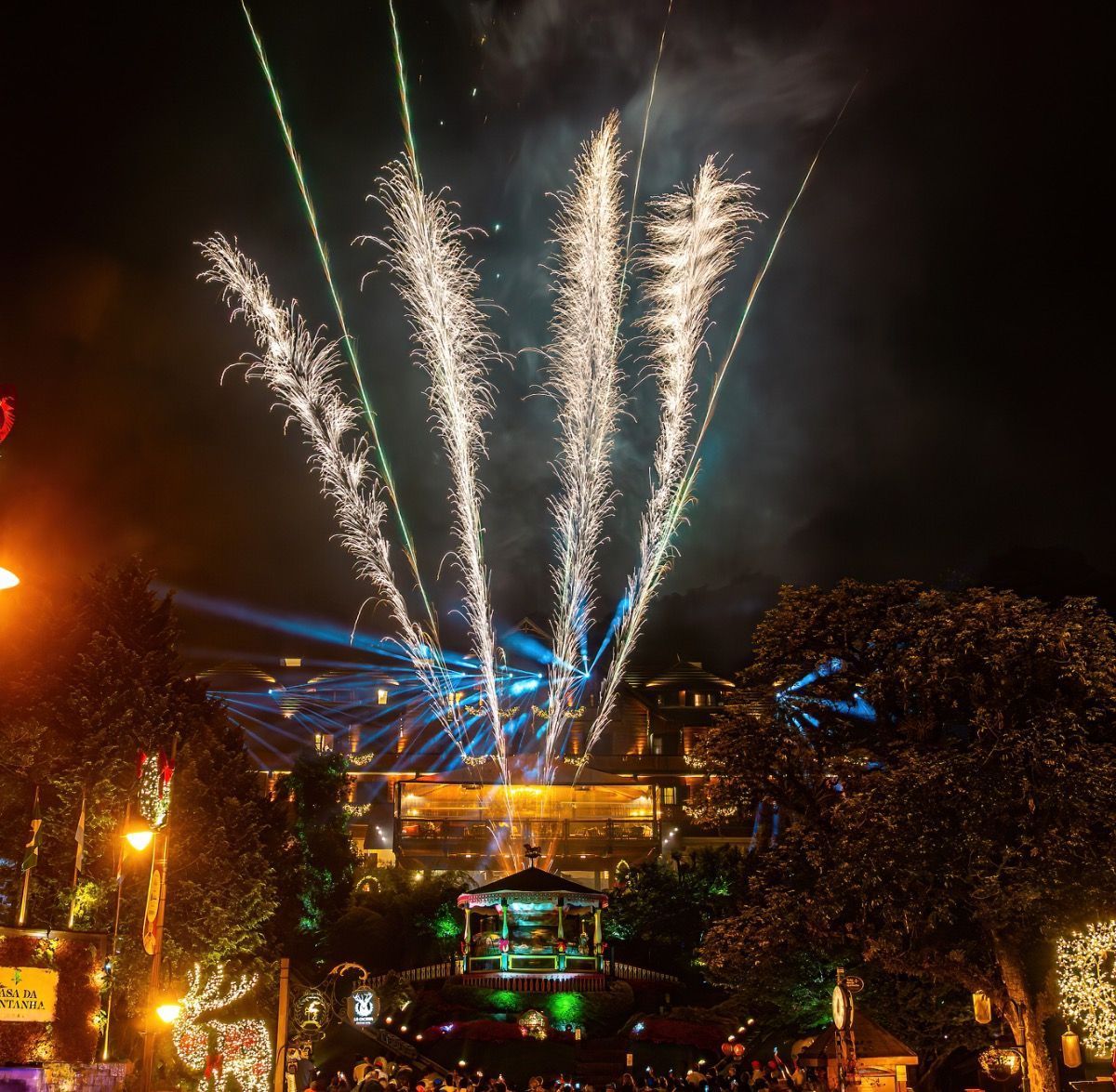Fogos de artifício explodem sobre um prédio iluminado à noite, deixando rastros azuis e brancos no céu.