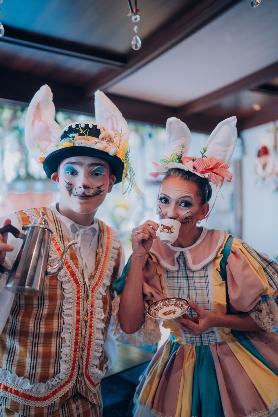 Duas crianças em trajes de carnaval coloridos e chapéus com penas, sorrindo dentro de casa.