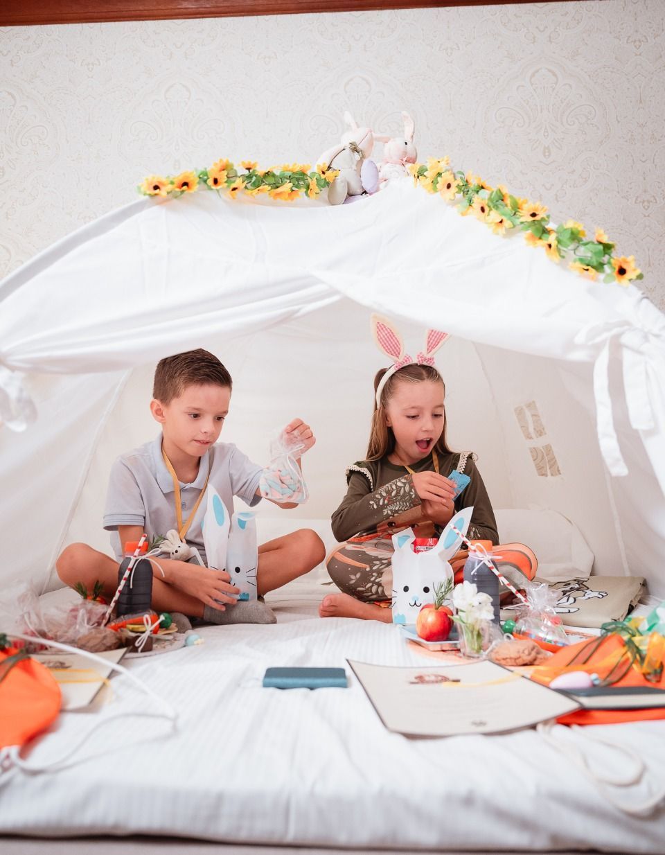 Duas crianças brincam dentro de uma tenda branca, cercadas por brinquedos e materiais de artesanato.