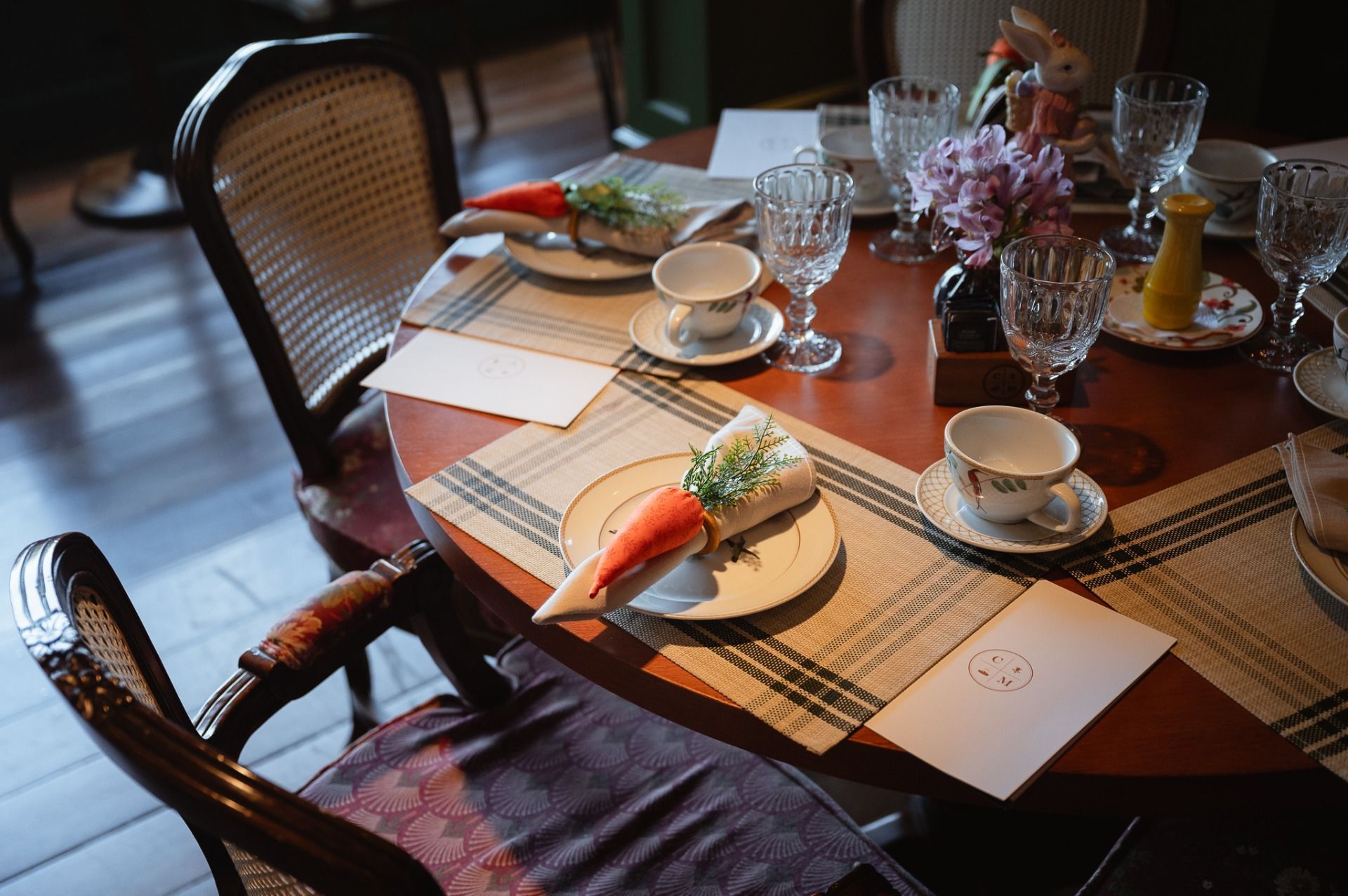 Prepare a mesa de jantar com jogos americanos de palha, xícaras, pratos e um arranjo de flores junto à janela.