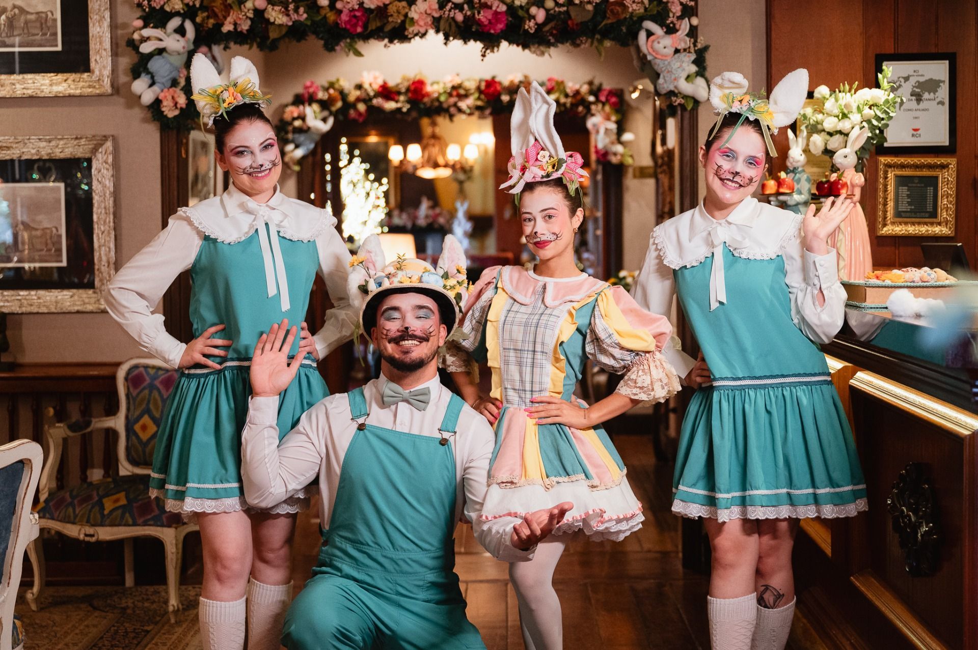 Quatro pessoas vestidas com trajes folclóricos em tons de verde-azulado e branco posando em uma sala repleta de flores.