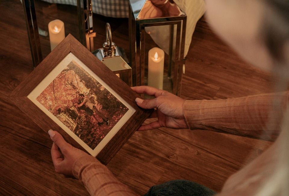 Mãos segurando uma obra de arte emoldurada sobre um piso de madeira ao lado de velas acesas e uma cadeira.