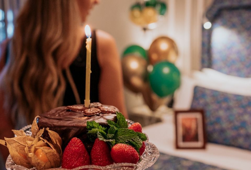 Bolo de aniversário com vela acesa, morangos e balões ao fundo.