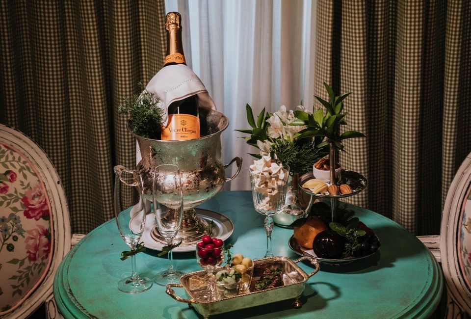 Mesa de sobremesas decorada com um balde de champanhe, flores, taças e doces sobre uma toalha de mesa azul-petróleo.