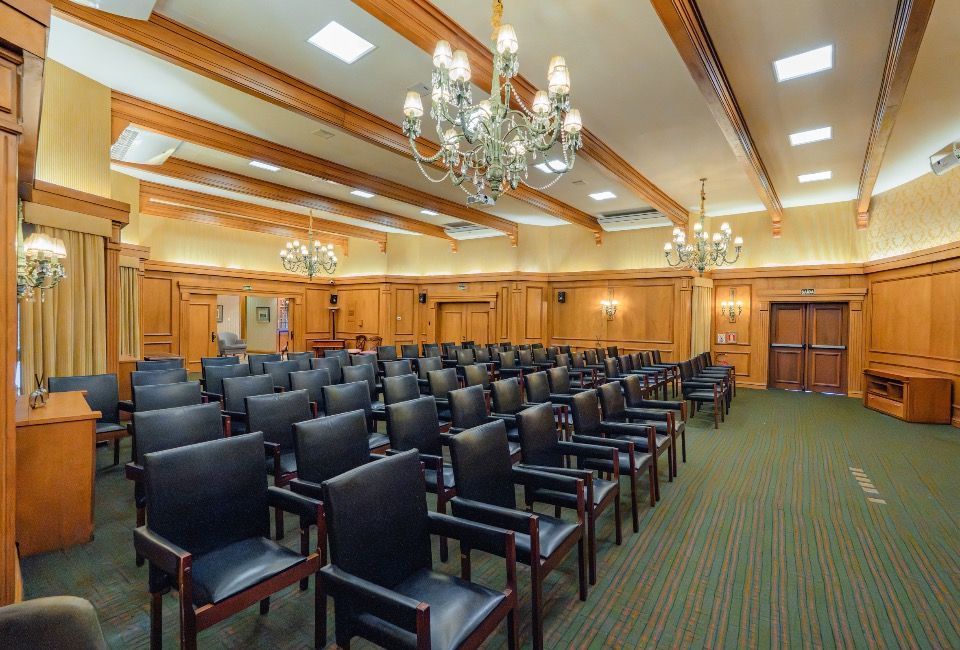 Sala de reuniões vazia, com painéis de madeira, fileiras de cadeiras escuras e iluminação de lustre.