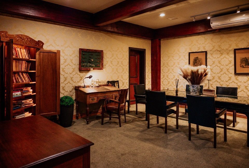 Sala de estar aconchegante com móveis de madeira escura, mesa, cadeiras, estantes e paredes em tom bege quente.
