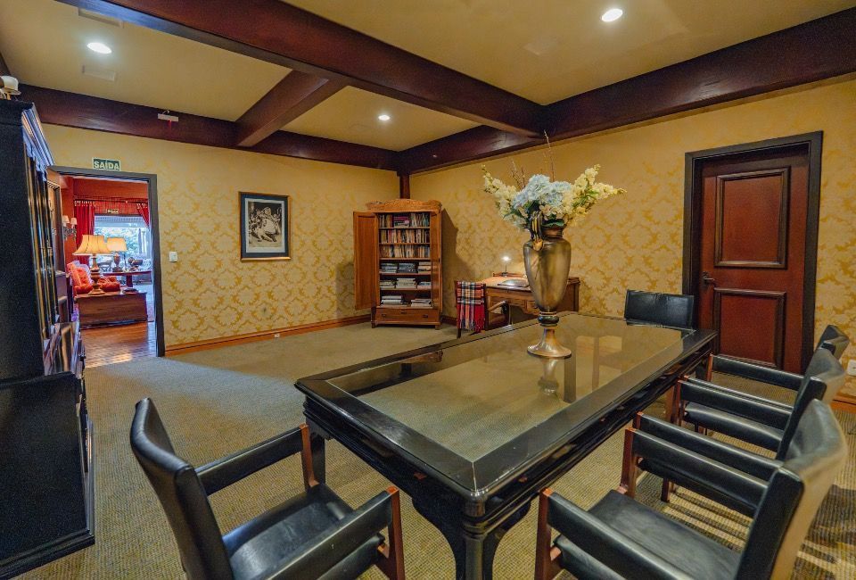 Sala de conferências com mesa de vidro, cadeiras pretas, detalhes em madeira e um vaso de flores em um suporte central.