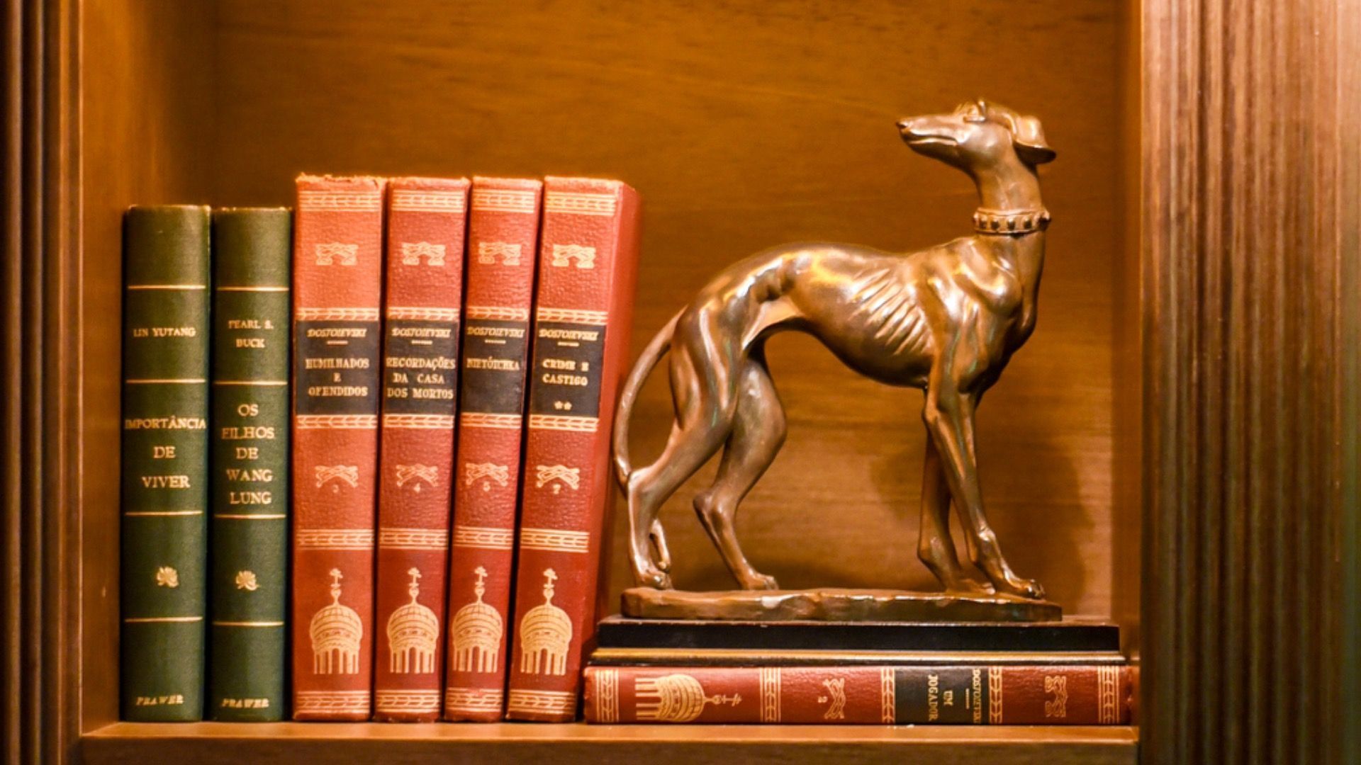 Estante de madeira com livros vermelhos e verdes e uma estatueta de bronze de um cervo em exposição.