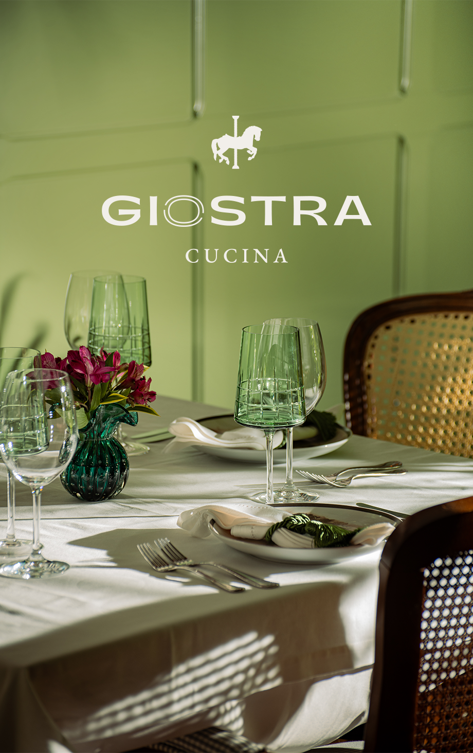 Elegante mesa de jantar posta na Giostra Cucina com copos verdes e luz solar aconchegante.