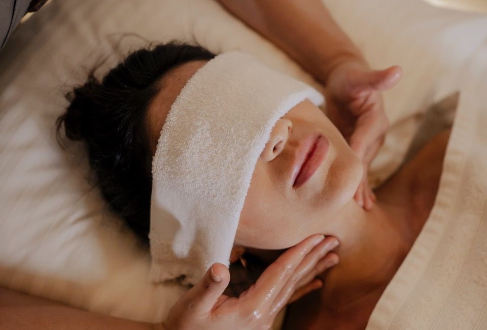 Tratamento facial de spa com uma mulher deitada, olhos cobertos por uma toalha, enquanto mãos massageiam seu rosto.