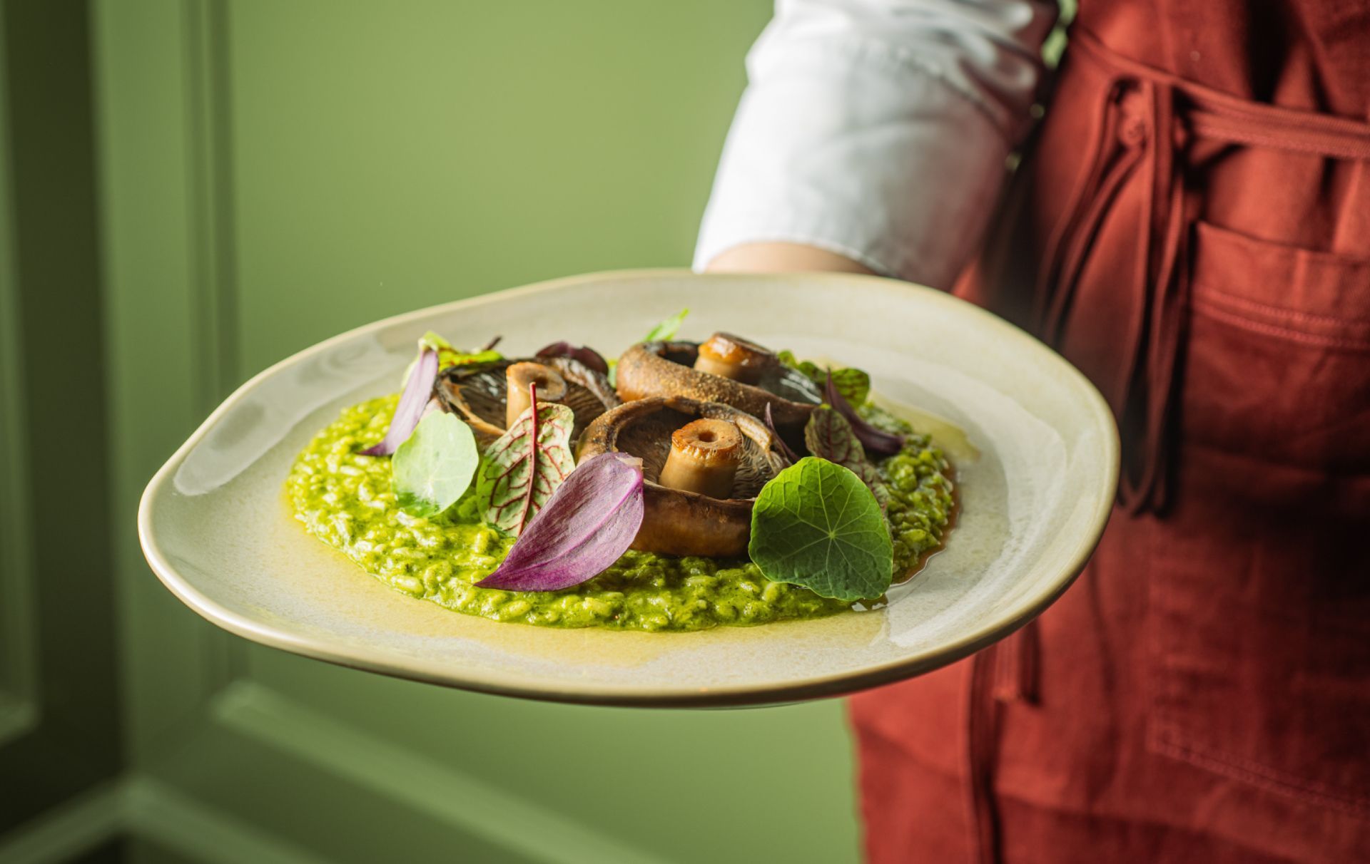 Prato gourmet com cogumelos e verduras, servido por uma pessoa com avental vermelho.
