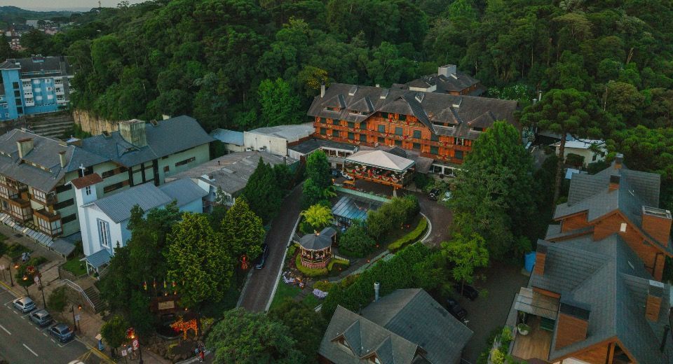 Vista aérea de um resort na encosta de uma colina, com edifícios alaranjados, rodeado por árvores e estradas sinuosas.