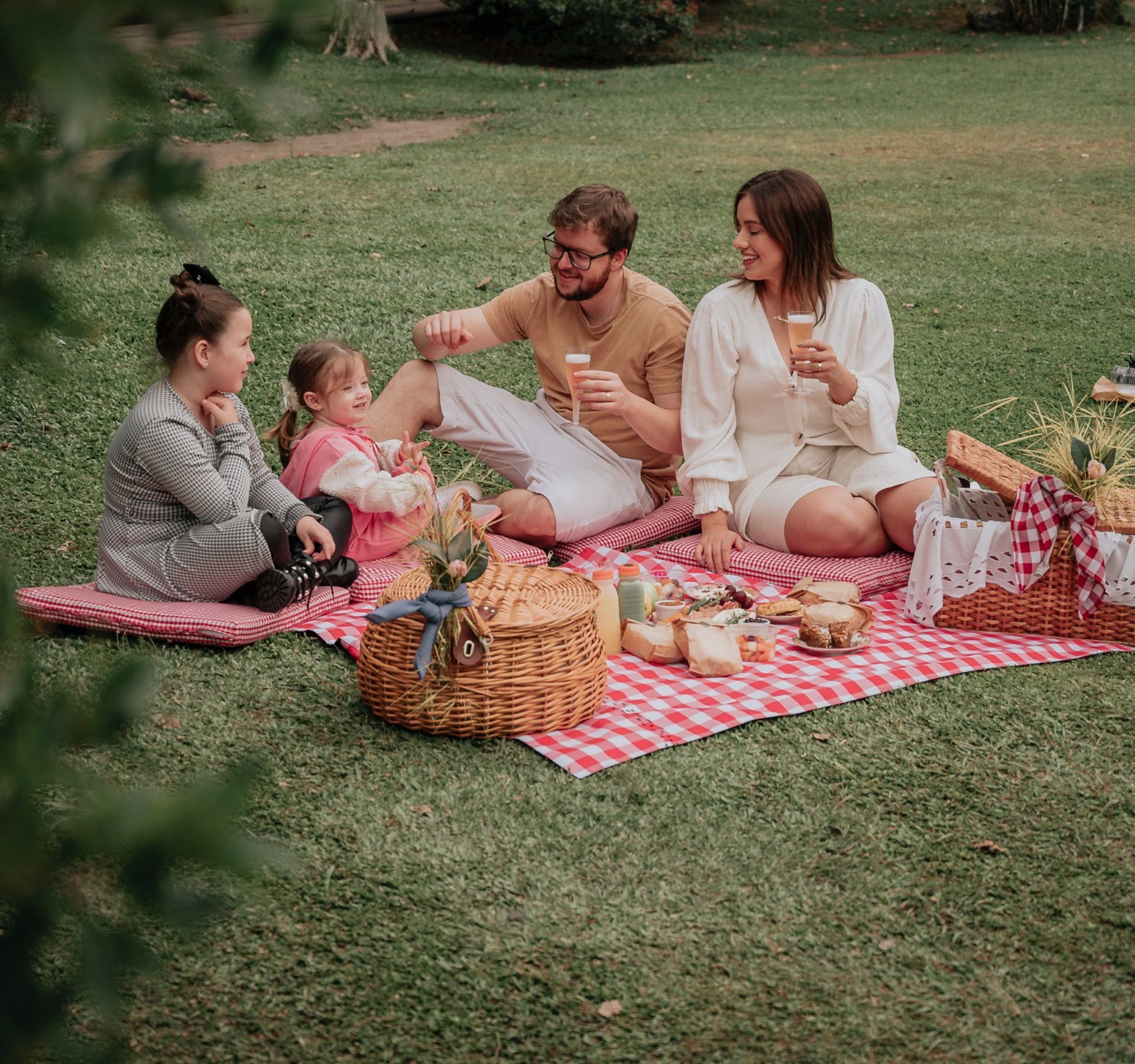 Família fazendo um piquenique em uma manta xadrez rosa em um parque gramado, compartilhando comida e bebidas.