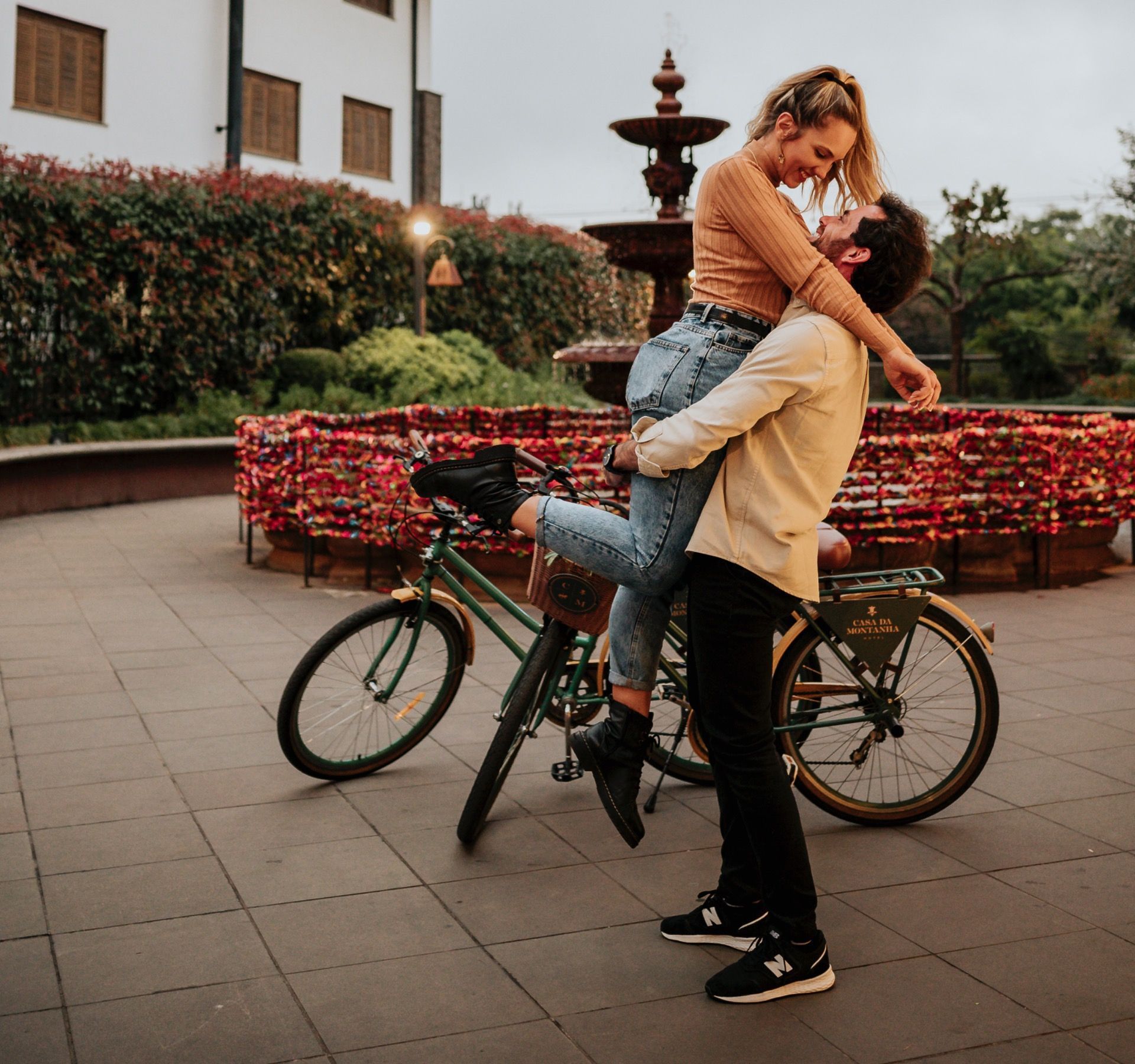 Duas pessoas se abraçando ao lado de uma bicicleta perto de um canteiro de flores e uma fonte ao ar livre.