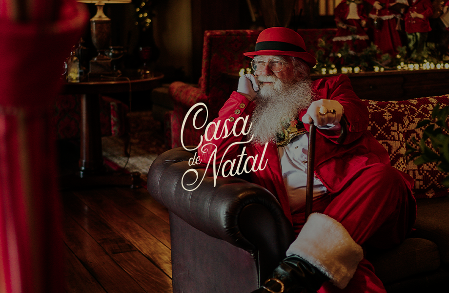 Papai Noel, vestido com um traje vermelho, está sentado em uma cadeira ao lado de um saco de brinquedos em um ambiente natalino aconchegante.