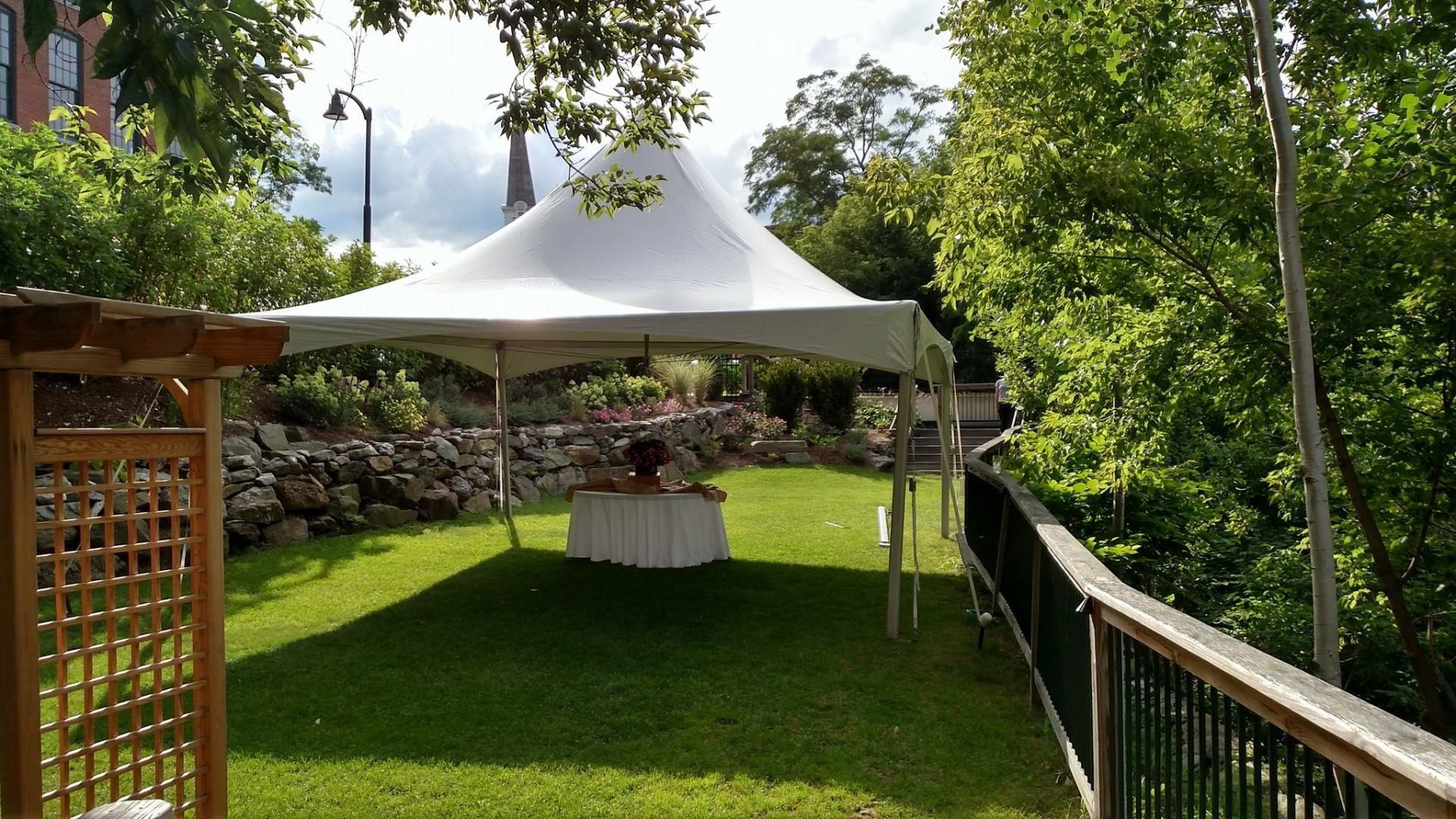 White canopy tent on a green lawn beside a wooden deck and stone wall, surrounded by trees.