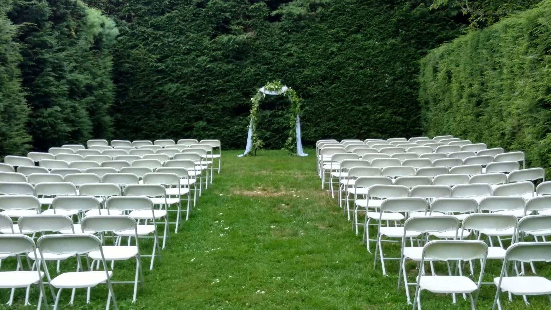 Rows of white chairs on a grassy aisle leading to a floral arch in a hedge-lined garden setting