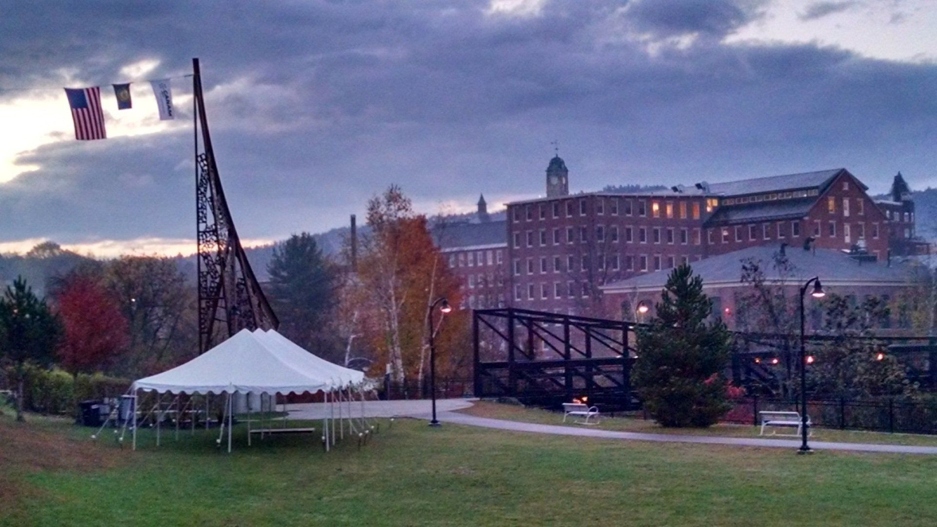 Tent-like structure on a grassy campus lawn at dusk, with a building and flags in the background