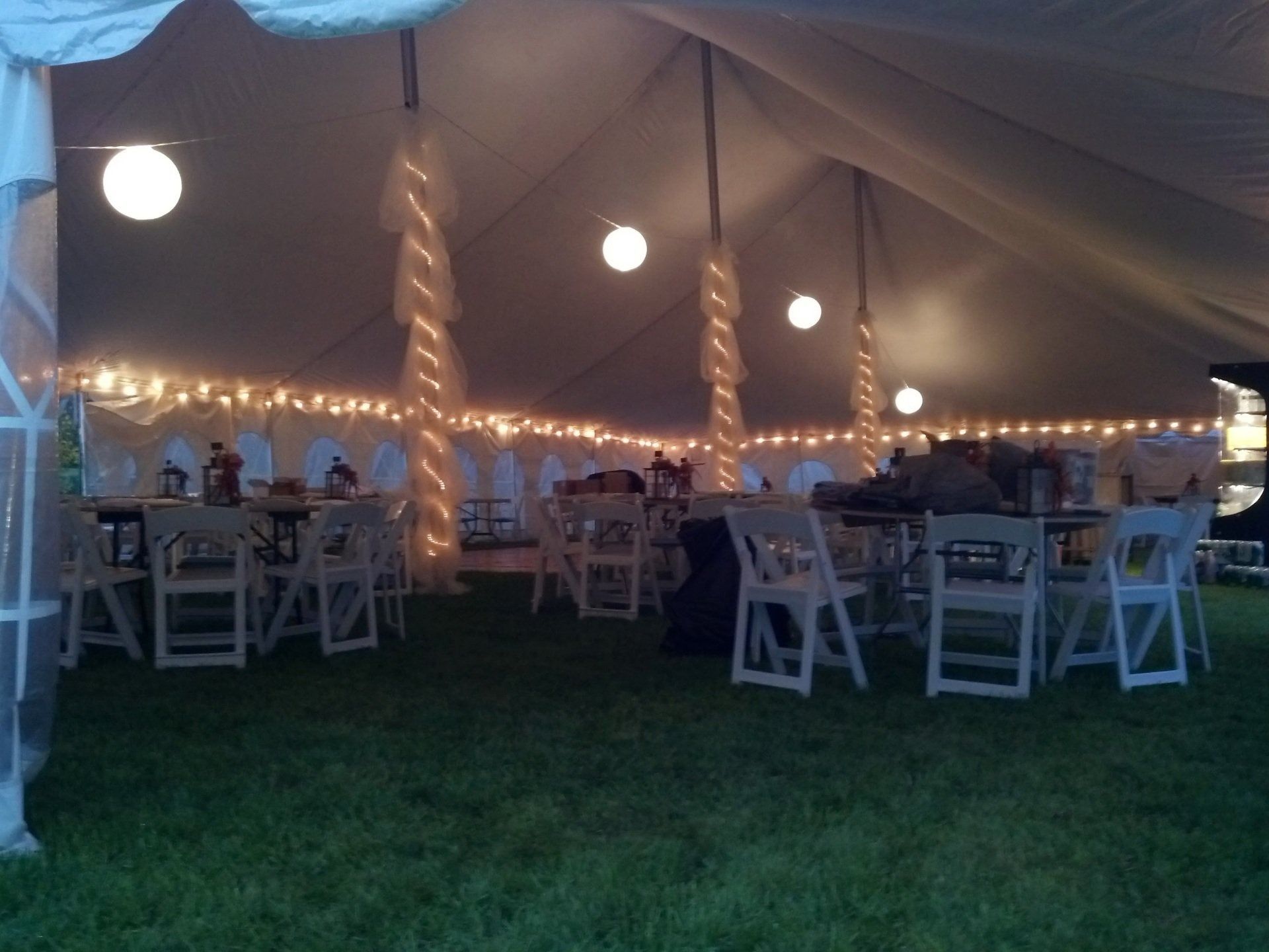 Tent-lit outdoor dining tent with string lights, white chairs, and tables on grass at dusk