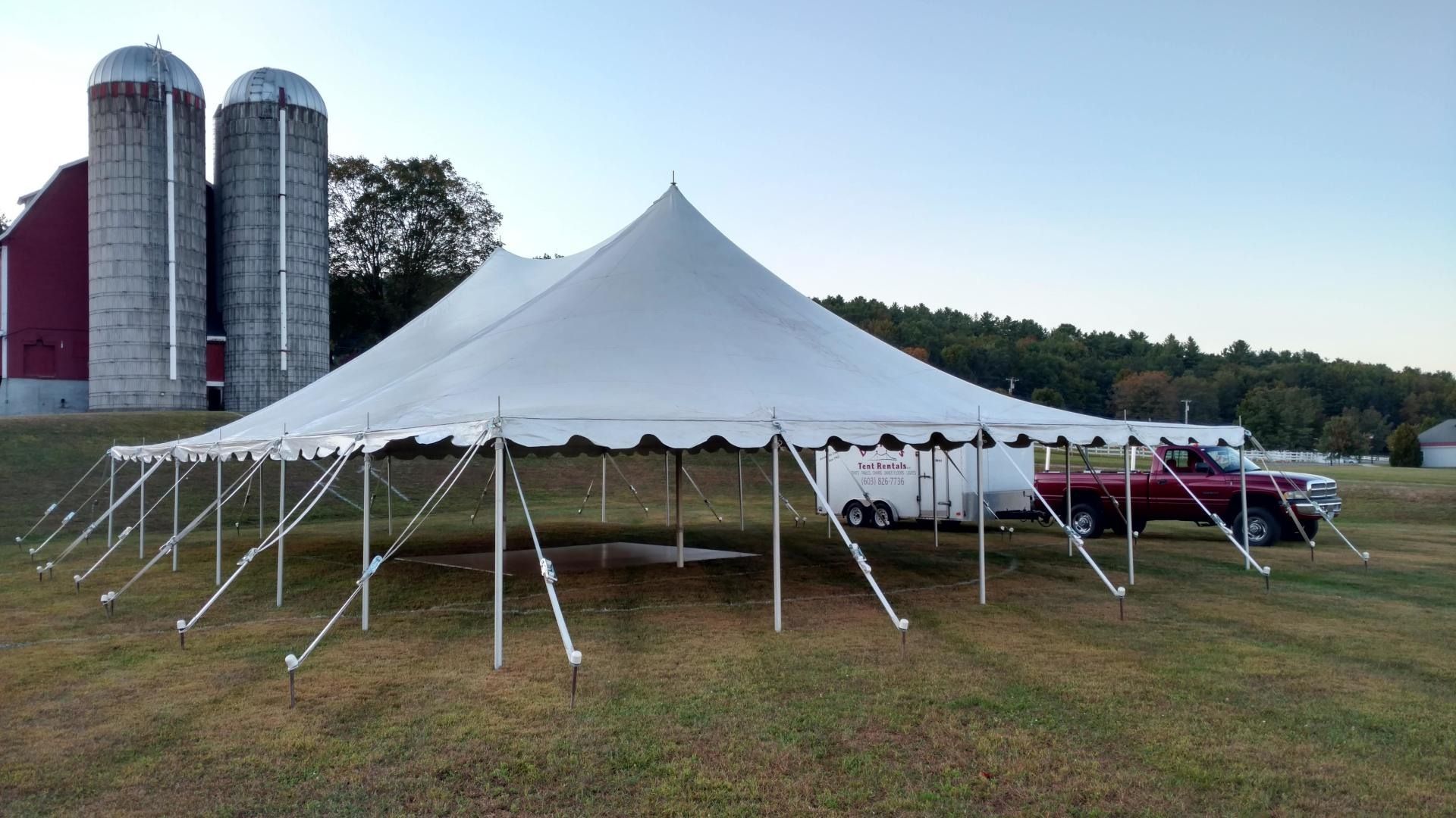 White event tent on a grassy field beside silos, with tables and cars in the background.