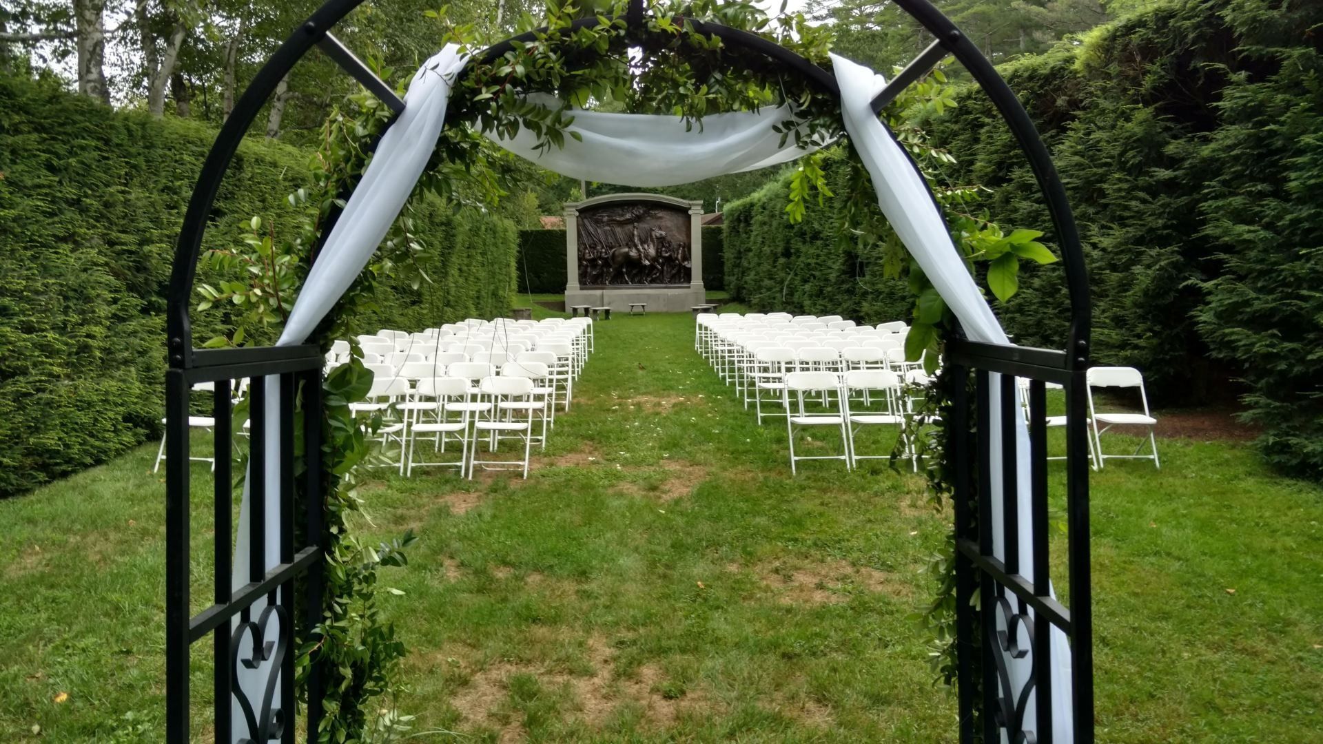 White wedding chairs under a draped garden arch on a grassy outdoor aisle