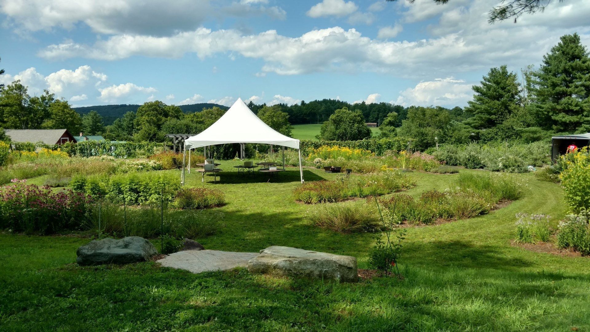 Garden with a white canopy tent, lush flower beds, rocks, and a cloudy blue sky.