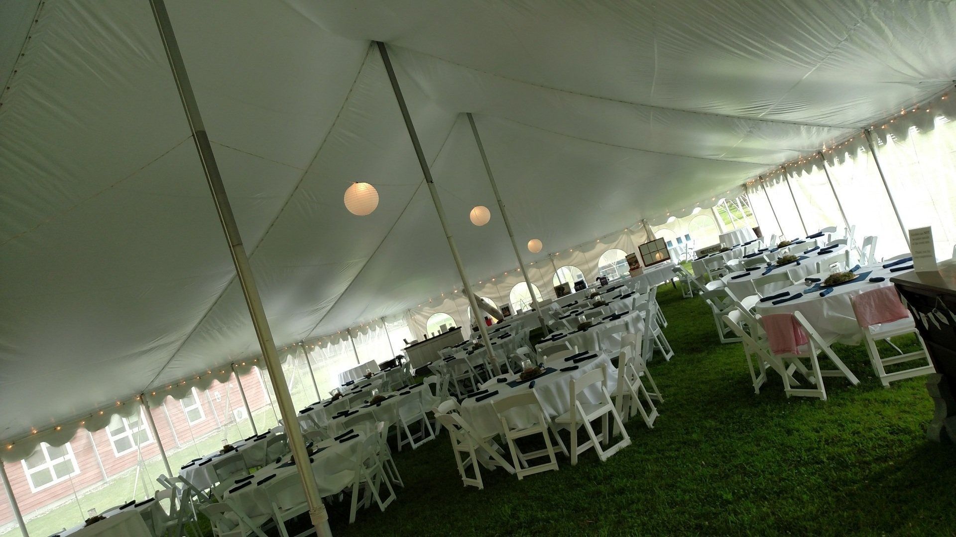 Inside a white event tent, rows of white chairs and round tables are set up on grass.