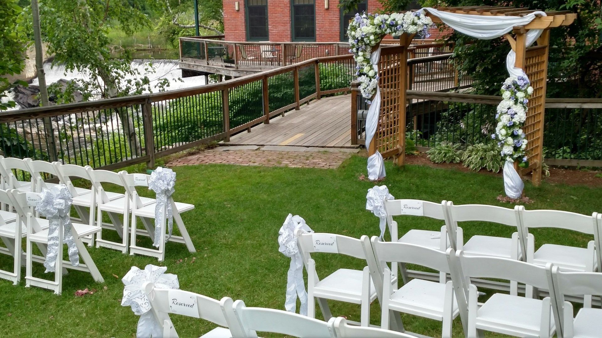 White chairs arranged on a grassy lawn for an outdoor wedding ceremony under a floral arch.