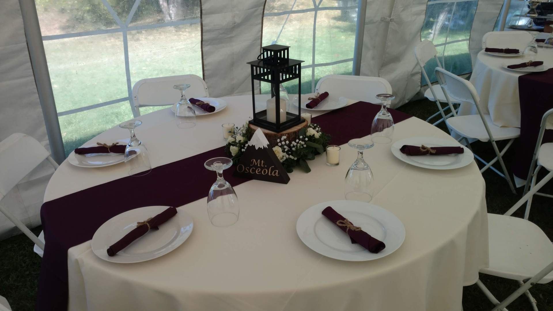Round banquet table with white and burgundy linens, candles, and folded napkins in a tented event setting