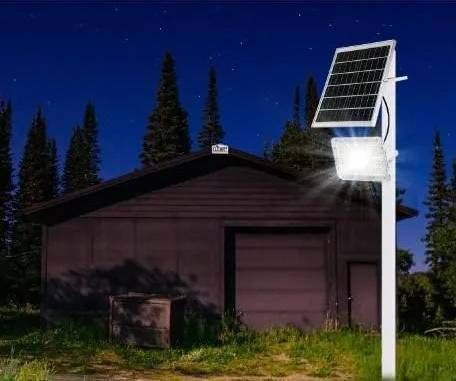 Una potente luz de seguridad LED montada en un poste con un panel solar, que ilumina el lateral de un edificio oscuro por la noche.