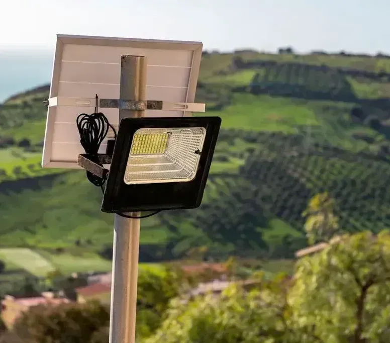 Un reflector LED alimentado por energía solar, montado sobre un poste, con un paisaje de colinas verdes y una costa lejana como telón de fondo.
