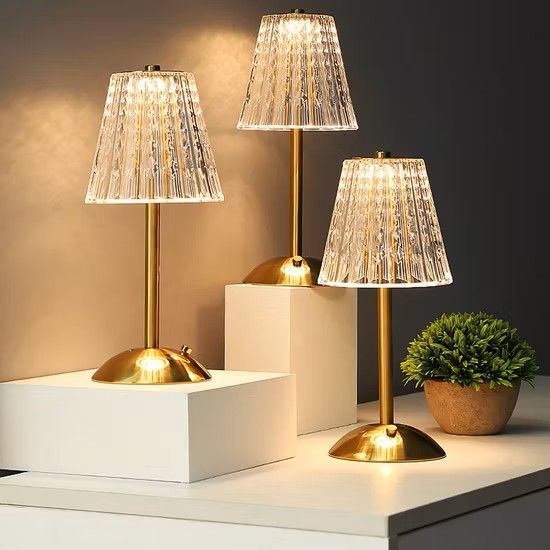 Tres elegantes lámparas de mesa con bases doradas y pantallas de cristal texturizado descansan sobre plataformas blancas junto a una pequeña planta en maceta.