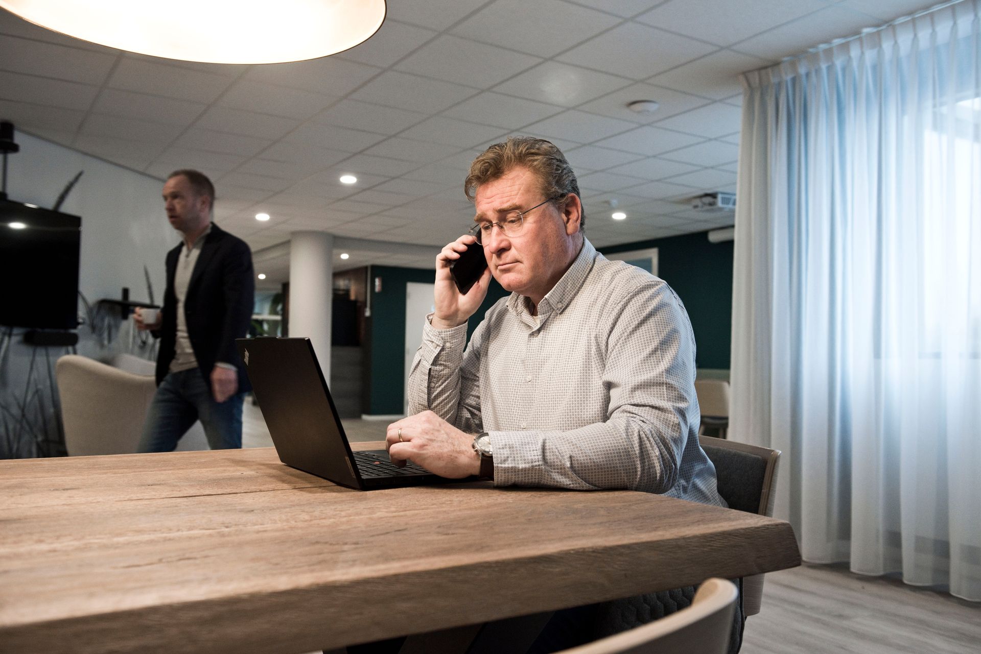 Two men at a table, looking at a laptop. The man on the left gestures, speaking. Neutral office setting.