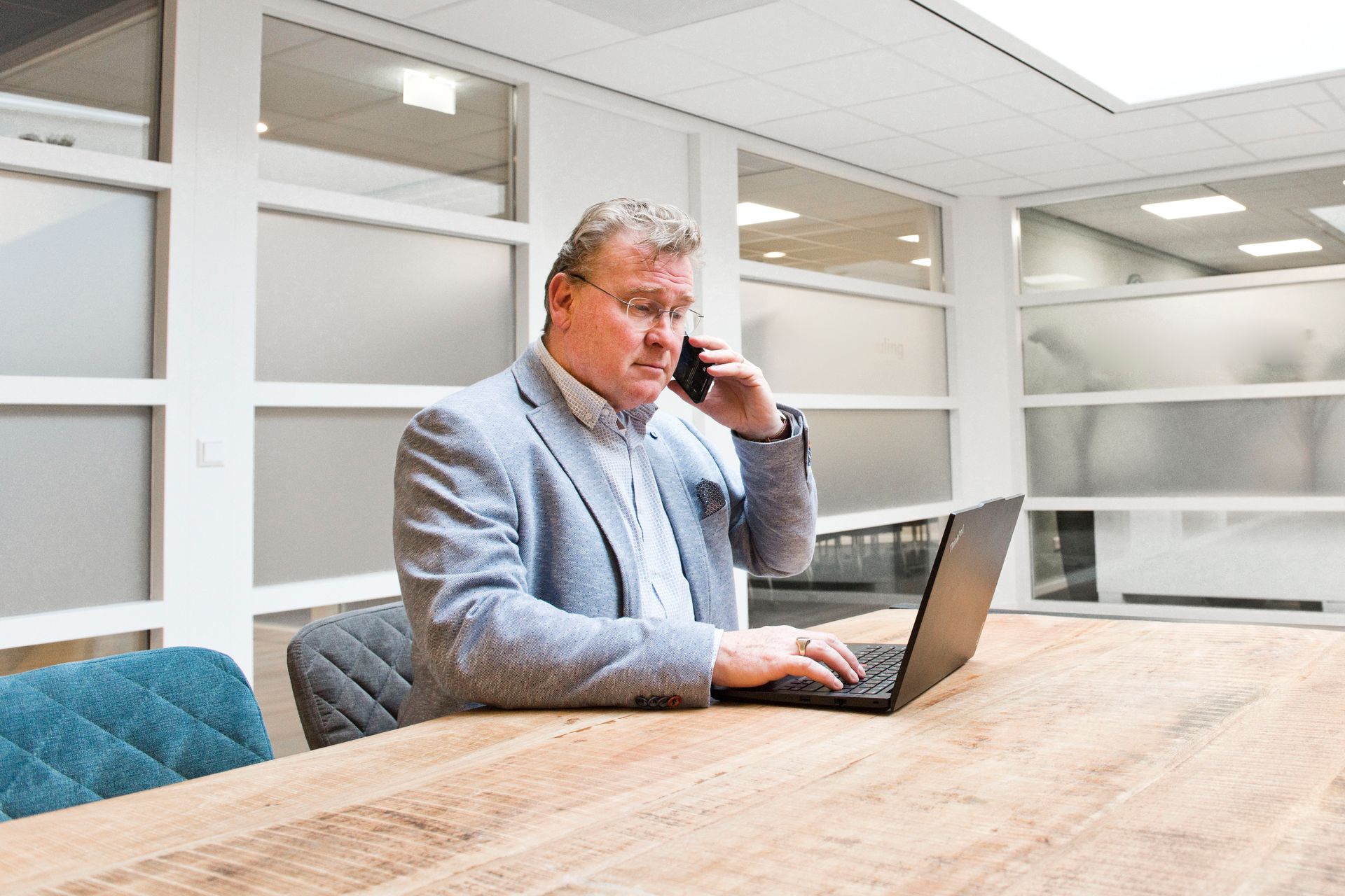 Two men at a table, looking at a laptop. The man on the left gestures, speaking. Neutral office setting.