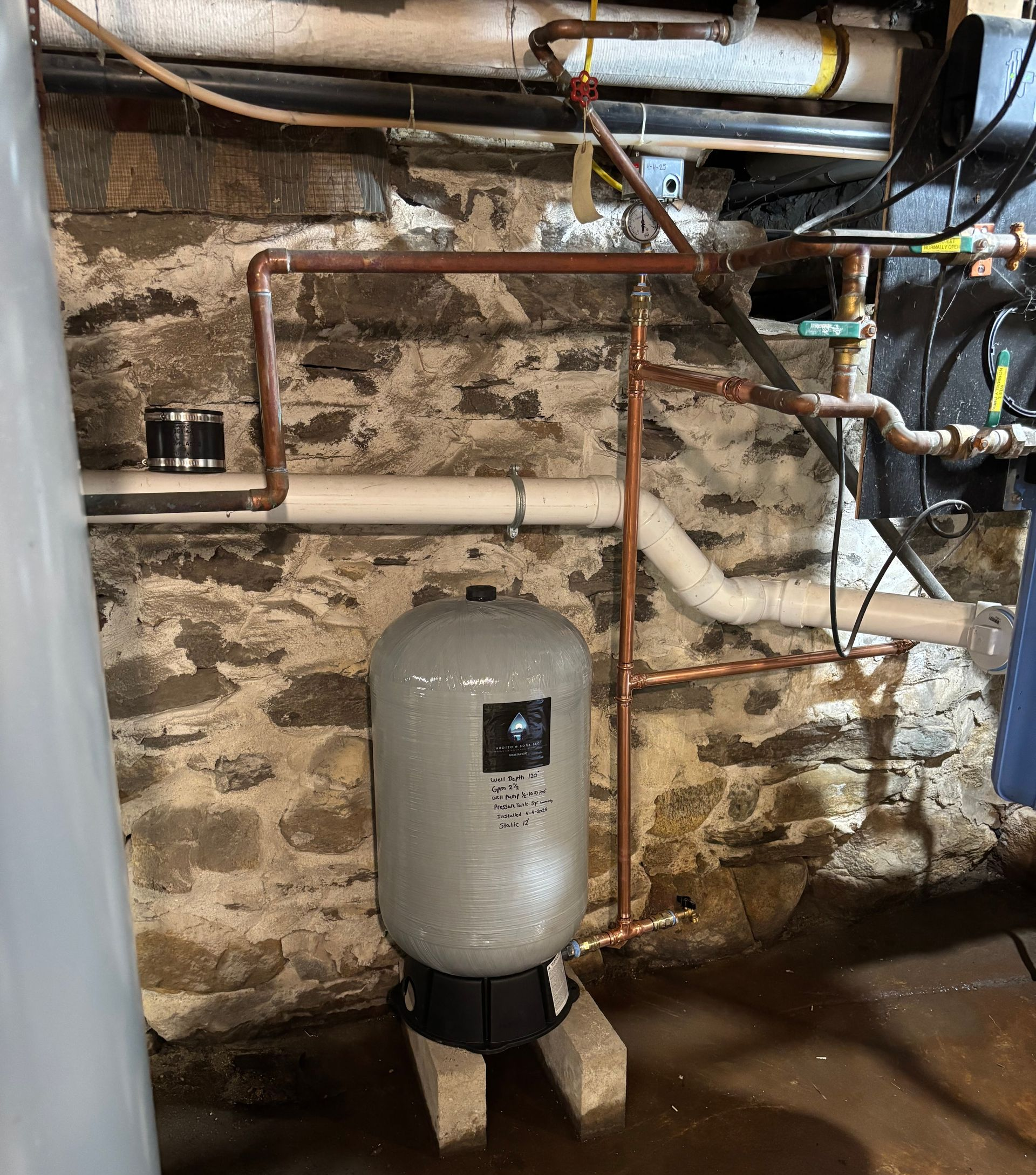 Plumbing in a basement with a pressure tank, copper pipes, and stone walls.