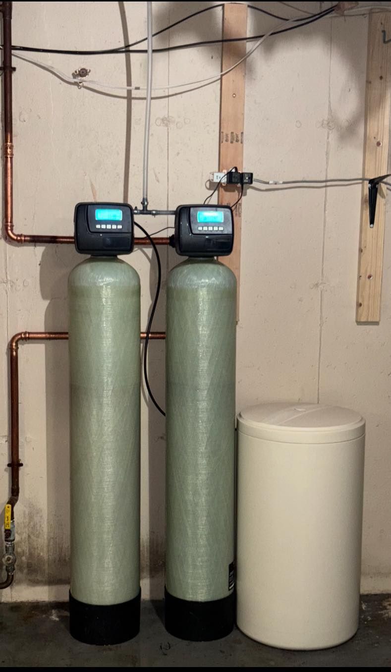 Two water filtration tanks with digital displays, beside a salt tank, against a cement wall. Copper piping visible.