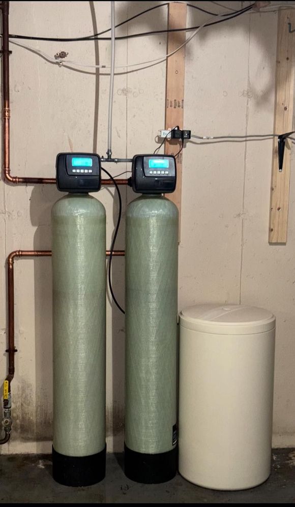 Two green water filtration tanks and a white brine tank in a basement setting.