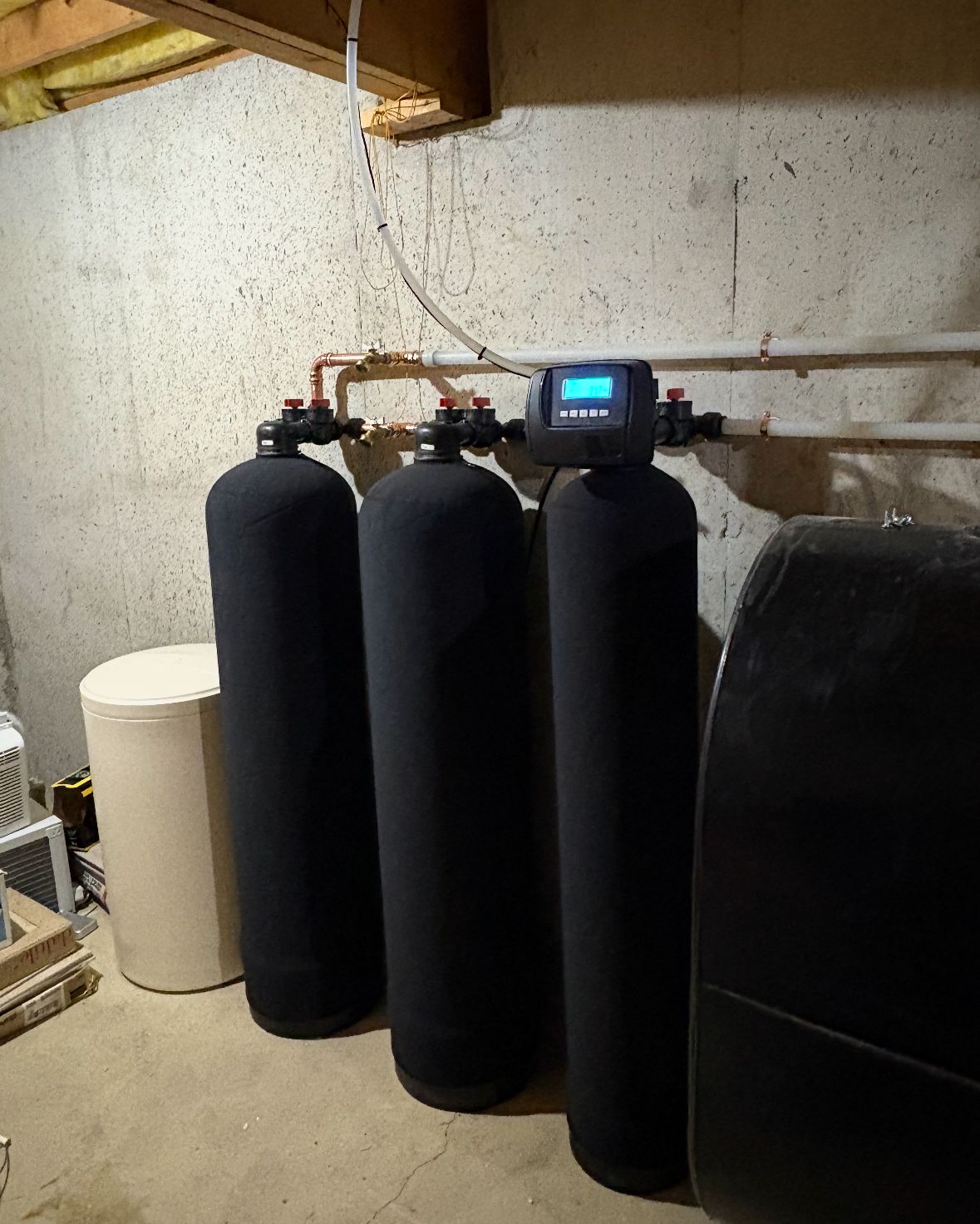 Water filtration system with three black tanks, a control panel, and a beige water heater in a basement.