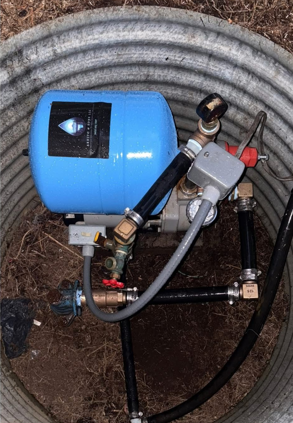 Blue water pressure tank with connected pipes, pressure switch, and valves inside a corrugated metal well.