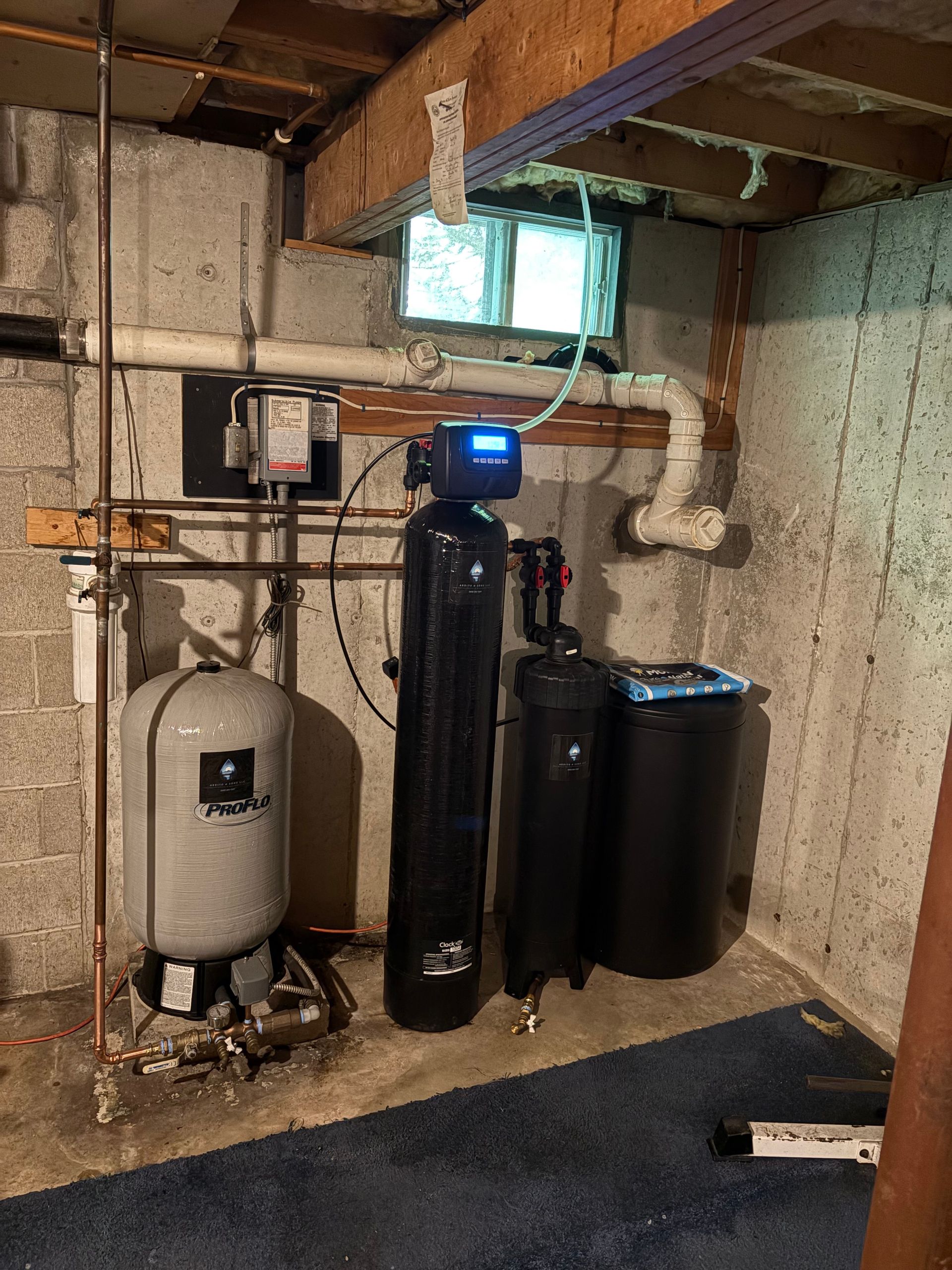 Basement water treatment system: pressure tank, softener, filter, and brine tank against concrete walls.