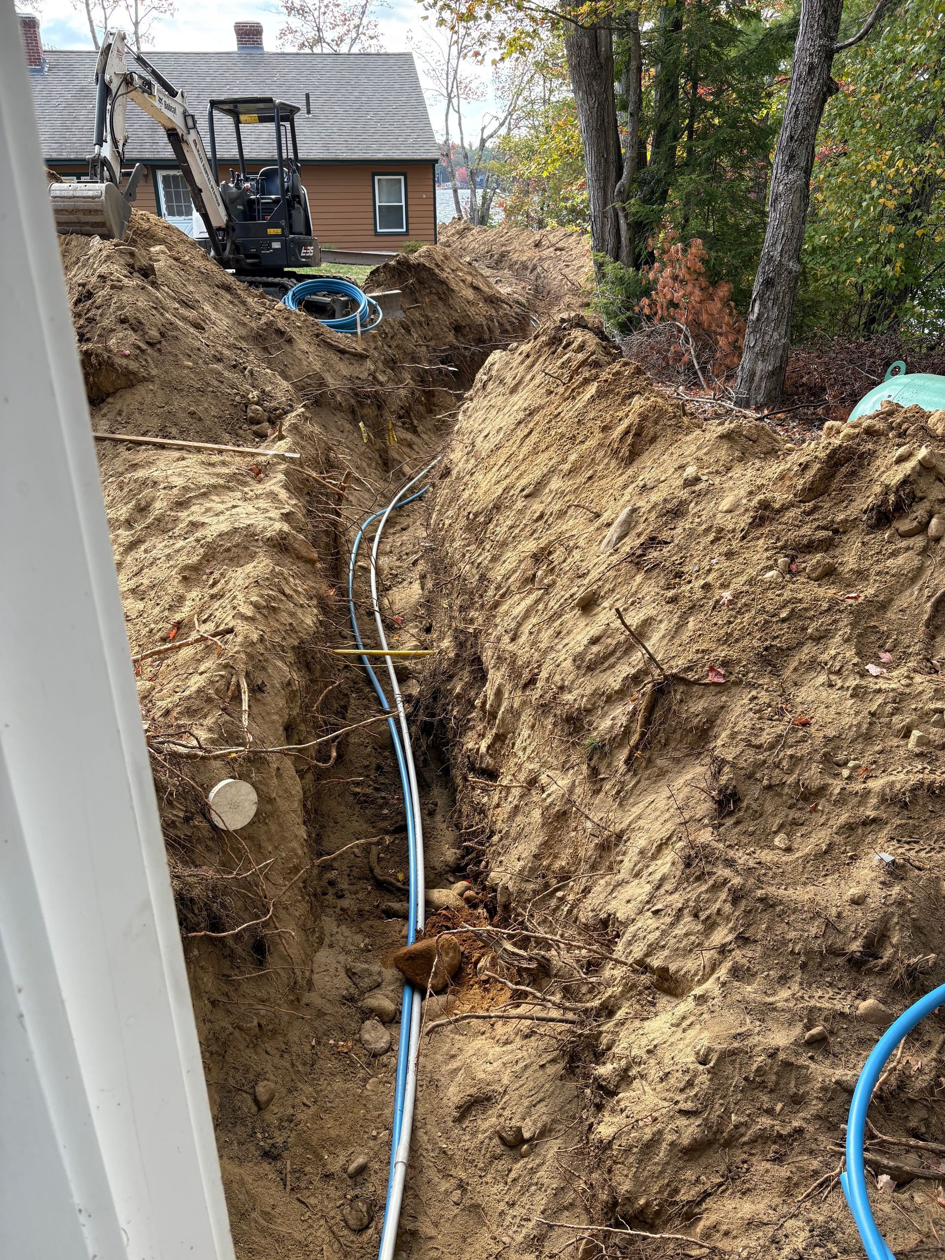 Trench with blue pipes for underground installation; mini-excavator in background; brown dirt; outdoor setting.