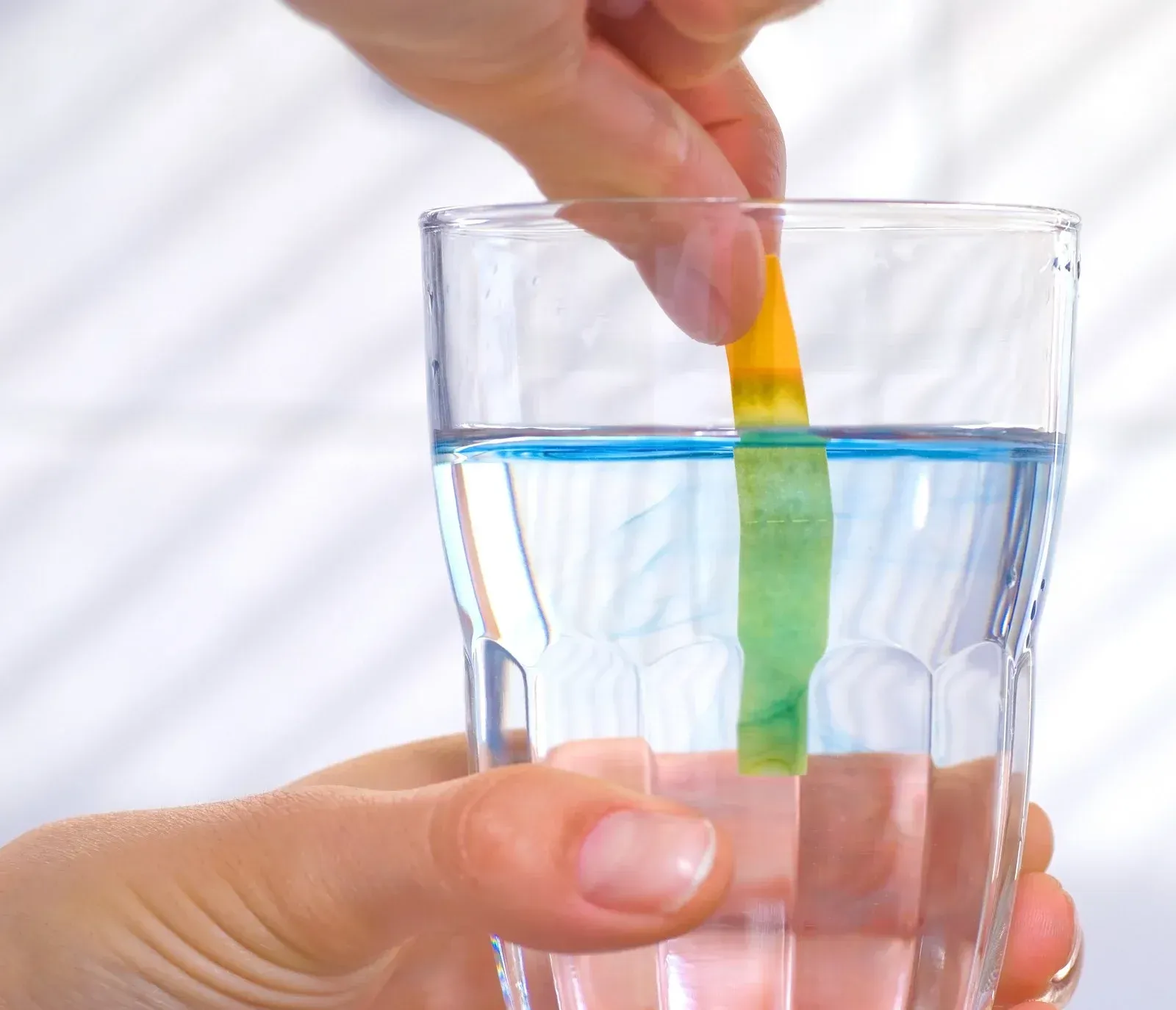 A hand holding a pH test strip in a glass of blue liquid; strip turns green.
