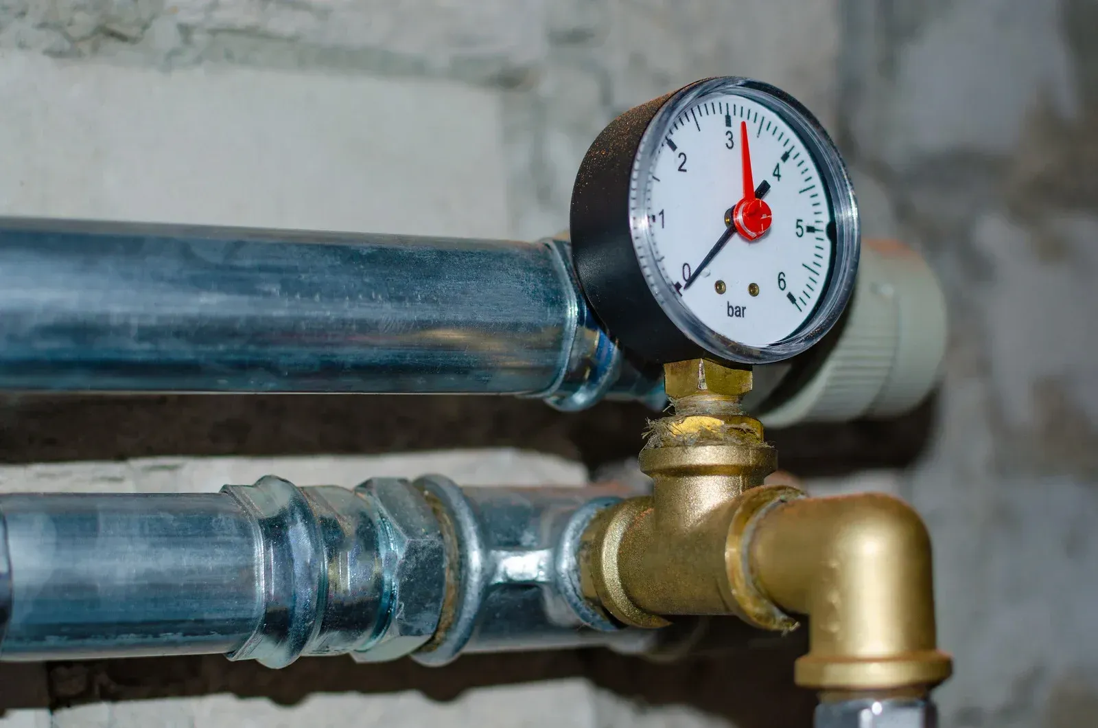 Pressure gauge on a brass fitting attached to two silver pipes. The gauge reads near 12.
