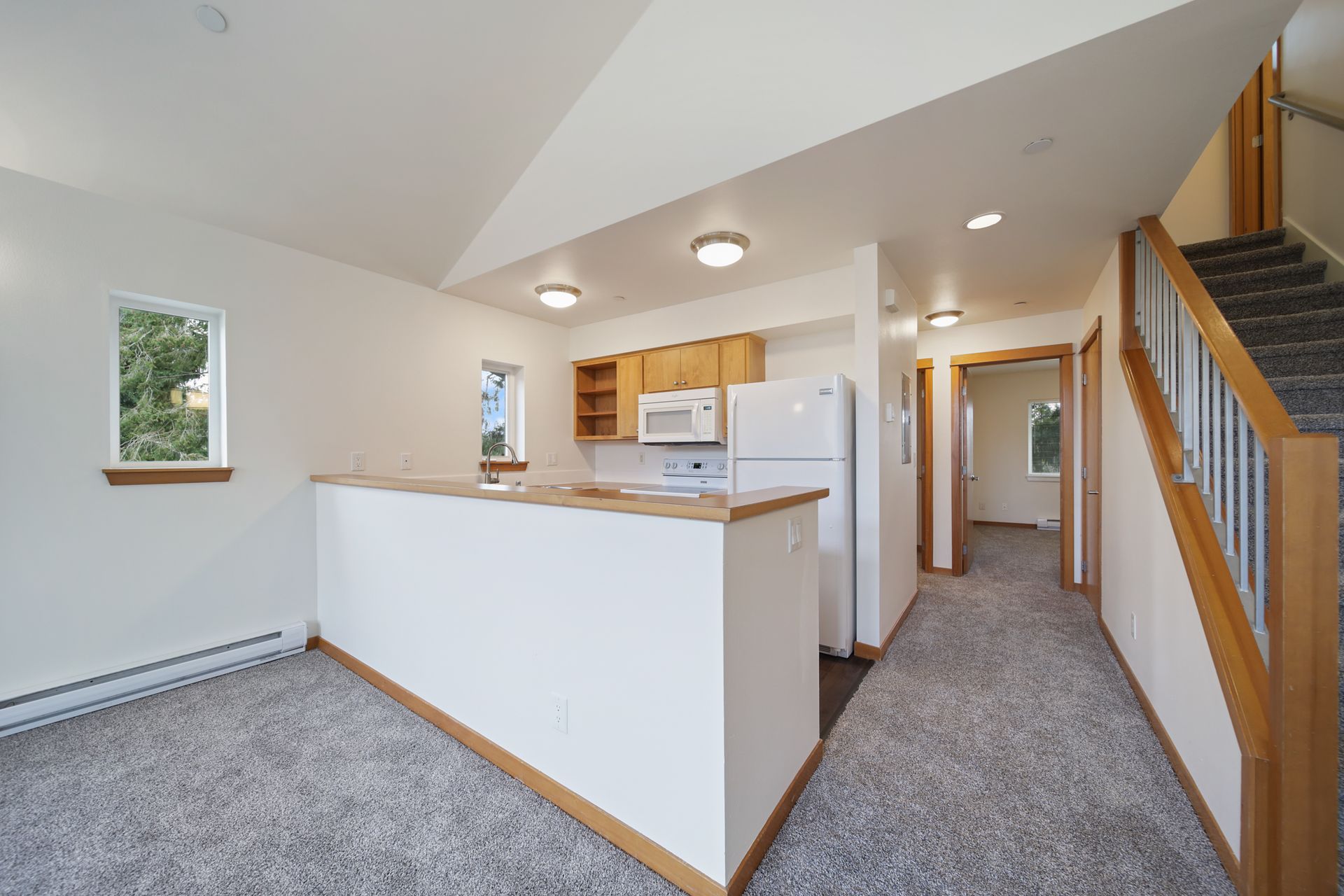 Bright open kitchen and hallway with carpeted floor, white walls, wooden trim, and stairway railing