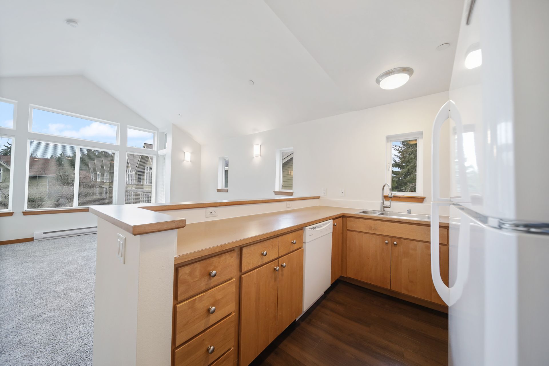 Bright modern kitchen with wood cabinets, white counters, and a view into a carpeted living area.